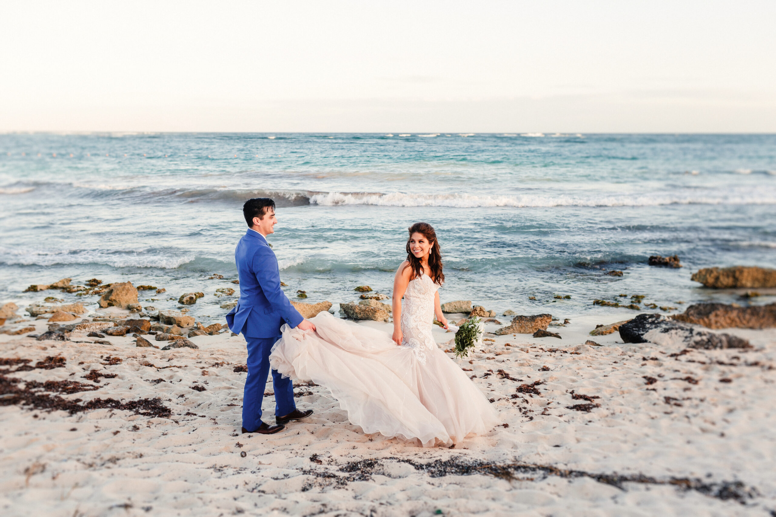 Mexico Elopement Packages - Bride and groom standing on a beach with ocean waves in the background, holding hands and looking at each other.